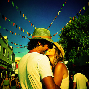 Colin Farrell and Kim Bordenave embrace on a vibrant city street festival. Bright sunlight, Colin wears a wide-brimmed hat, Kim in a casual white t-shirt.