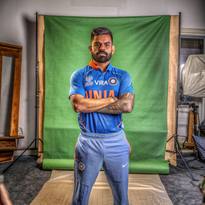 Virat Kohli, cricket star, stands alone on a green screen. Serious expression. Blue jersey with '10' and 'VIRAT' visible. Bearded face.