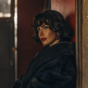 Mia Kirshner, with dark, voluminous bob hairstyle and bold red lipstick, poses against a red backdrop. She wears a luxurious black coat and red earrings, capturing attention with her serious expression.
