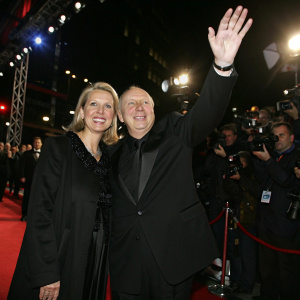 Caucasian couple, Reiner Calmund and Sylvia Calmund, dressed in black, wave at celebrity event. Bright lights and cameras.
