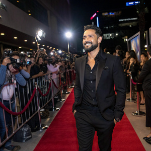Juan Ugarte, at the launch of his new movie 'Casi Treinta'. The scene captures him in a sharp black suit, with a well-groomed beard. Against a backdrop of flashing cameras and excited fans on a bustling red carpet event.