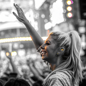 Victoria Justice, blonde hair in ponytail, waving at fans. Bright neon lights and crowd in the background.