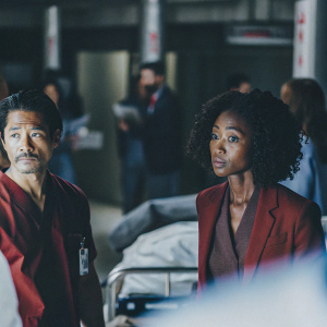 Yaya DaCosta and Brian Tee in a tense scene from 'Chicago Med'. Intensive care unit setting, with blurred medical equipment and dim lighting. Both actors in formal attire, Yaya in blue scrubs, Brian in red. Their expressions convey seriousness.
