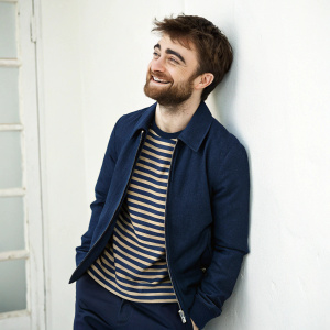 Daniel Radcliffe, smiling with a beard, dressed in a casual navy jacket and striped shirt. Solo artist portrait against plain white wall