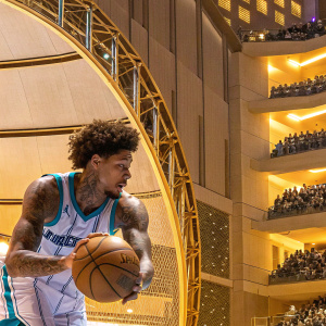 Kelly Oubre Jr., dynamic pose, dribbling basketball, in teal and white Hornets jersey. African descent, curly hair, tattoos. Crowded arena in background.