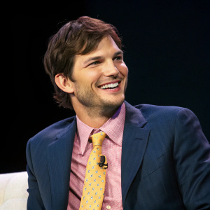 Ashton Kutcher, smiling at camera. Attending tech conference. Wears navy suit, pink shirt, yellow tie. Dark background