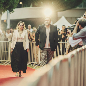 Vincent D'Onofrio and Carin Van Der Donk attending a film festival premiere. D'Onofrio in a dark suit with a beard. Carin in a black skirt and white blazer. Crowded red carpet with flashing cameras.