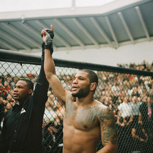 Abel Trujillo, a mixed-ethnicity UFC fighter, celebrating his win. Shirtless with visible tattoos, raising his arm. Staff member of mixed ethnicity stands beside him. Energetic crowd in background