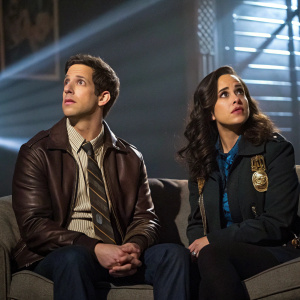 Andy Samberg and Melissa Fumero on the set of Brooklyn Nine Nine. Authentic costumes, they sit on couch in dramatic lighting.