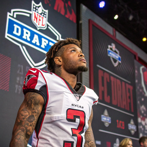 Football player with tattoos, intense expression, wearing Atlanta Falcons jersey. NFL Draft event, surrounded by NFL Draft branding and logos.