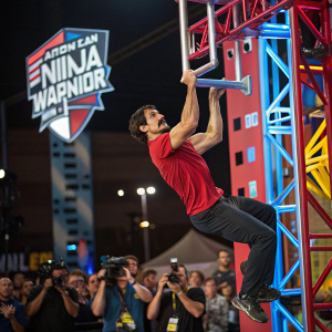 Man with mustache performing daring obstacle course at American Ninja Warrior Semifinals. Climbing vibrant ninja-themed structure in red shirt and dark pants. Crowd and photographers capture the moment against backdrop of event logo.