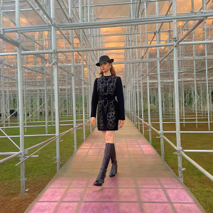 Sophie Turner at fashion gala. Wearing black fitted dress with intricate embroidery, matching hat, knee-high boots. White scaffolding and pinkish-purple flooring in the background. Confident pose.