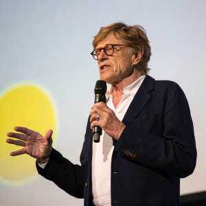 Robert Redford, an older white male with glasses, addressing an audience at a speaking event. He wears a dark blazer over a white shirt and actively gestures with his hands. The backdrop is plain and light-colored with a bright yellowish light source.