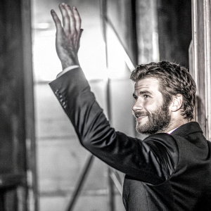 Liam Hemsworth, actor, with a beard, black suit. Hand raised, smiling slightly. In a dimly lit venue, the background is blurred.
