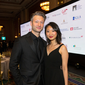 Peter Scanavino and Lisha Bai attend a gala event. Peter in a black suit, Lisha in a black dress. They pose with sponsors logos displayed on a large screen behind them.