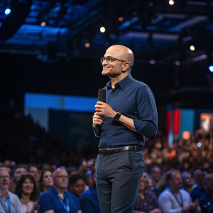 Satya Nadella, the CEO, is addressing a tech conference. He's dressed in a dark blue shirt and glasses. A bustling crowd of attendees fills the background.