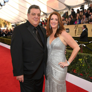Steve Schirripa and Laura Schirripa at the SAG Awards. Steve in a black suit, Laura in a silver gown. Stars and paparazzi in the background.