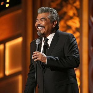 George Lopez, grinning broadly, center stage at the AFI Life Achievement Award gala. Wearing a black suit and tie, making an engaging speech to the audience. Warm, golden lighting