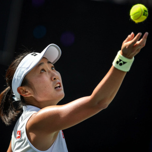 Misaki Doi, East Asian tennis player, in mid-swing. White visor and athletic wear. Dark background. Tennis ball mid-air. Intense focus on serve.