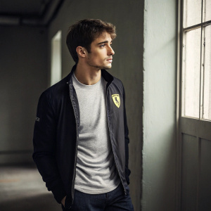 Charles Leclerc, Formula One driver, in a casual outfit. Grey t-shirt with Ferrari logo, black jacket. Standing alone in a dimly lit room.