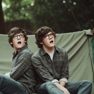 Niall Horan, close-up shot. Youthful face, wearing gray flannel shirt and jeans. Classic glasses, plain white background