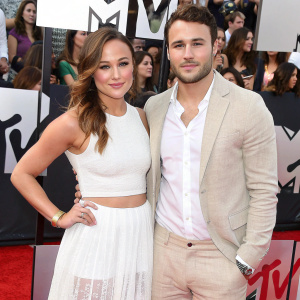 Briana Evigan and Ryan Guzman at the MTV Movie Awards. Red carpet event. MTV logo backdrop. Diverse crowd. Briana in crop top and white tiered skirt. Ryan in white shirt and beige structured blazer.