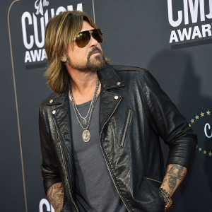 Billy Ray Cyrus, rugged look, at Country Music Awards. Black leather jacket, casual t-shirt, sunglasses. Tattoos visible. Dark, branded wall backdrop