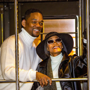 Will Smith and Jada Pinkett-Smith, an African American couple, posing for a magazine cover shoot. Black and white photography. Will in a white turtleneck, Jada in a black hat and sunglasses. Warm and friendly expressions.