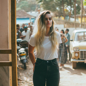 Alice Dellal, Caucasian female, long blonde hair in loose waves, standing in front of a bustling street scene. She wears a casual outfit, including a white t-shirt and black jeans.