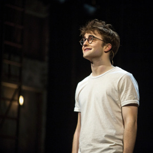 Daniel Radcliffe, young actor, wearing a simple white t-shirt. Messy hair and glasses, friendly expression. Standing alone on a dimly lit stage.