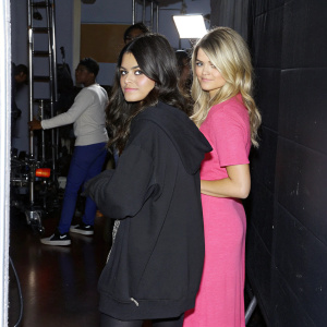 Sofia Black-D'Elia and Kaitlin Olson on the set of their TV show. Sofia in a casual black hoodie, Kaitlin in a bright pink dress. A bustling studio backdrop