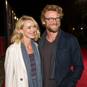 Naomi Watts and Simon Baker at a charity gala. Smiling, casual attire, dark red carpet background