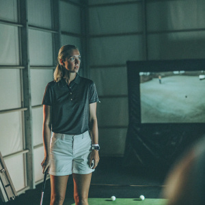 Jessica Korda, wearing a Nike polo shirt and white shorts, standing in front of a dimly lit indoor golf simulator. Focused look, surrounded by golf balls and clubs.