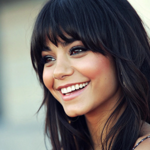 Vanessa Hudgens, with vibrant smile, styled dark bangs, close-up shot. Faint blurry backdrop.