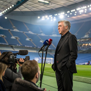 Carlo Ancelotti, dressed in a tailored black suit, addresses the media at the post-match press conference. Stadium backdrop, flashing cameras.