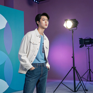 Digital portrait of Sehun from EXO. Wearing a casual outfit, standing in a modern art studio. The background is a gradient of purple and blue with abstract shapes. Bright studio lights illuminate his face.