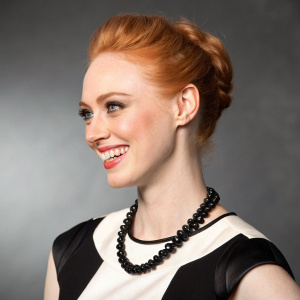 Close-up portrait of Deborah Ann Woll, white actress with vibrant red hair styled up. Wearing a stylish black and white ensemble with a black necklace. Shot against a gradient gray backdrop, capturing her radiant smile and upper body.