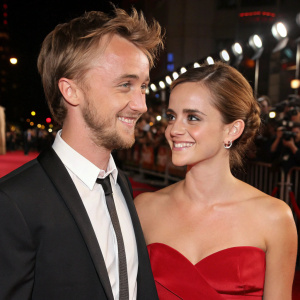 Tom Felton, with stubble and messy hair, Emma Watson in an elegant updo, smiling softly. Both at a glamorous movie premiere.