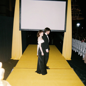 Rosemarie DeWitt and Jon Krasinski at a gala event. The venue is brightly lit with red carpet. They are standing beside a large screen showcasing awards