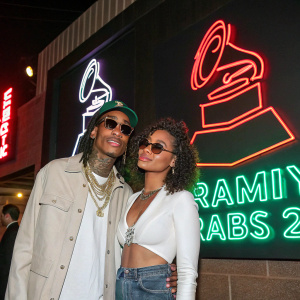 Wiz Khalifa and Izabela Guedes at the Grammy Awards after-party. Wearing casual attire, posing in front of large neon signage with sponsor logos.