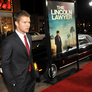 Ryan Phillippe, dressed in a black suit with a red tie, stands beside a movie poster for 'The Lincoln Lawyer'. The backdrop features a sleek car and vibrant promotional graphics. Taken inside an award show.