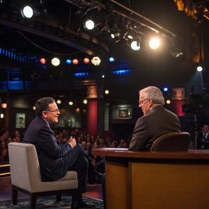 Stephen Colbert interviewing Bob Woodward. Studio audience present. Warm studio lights illuminate the scene.