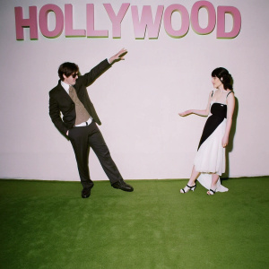 Steven Spielberg and Destry Allyn Spielberg at a Hollywood event. Dark suit and casual sweater for Steven, white and black dress for Destry. Backdrop features bold text and logos.