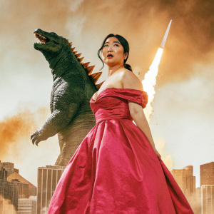 An Asian woman confidently stands in front of a colossal Godzilla, with a bustling futuristic cityscape and raging fire in the background. She wears a red off-the-shoulder gown. The scene is from the promotional material for the movie 'Godzilla vs Kong'.