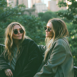 Ashley and Mary-Kate Olsen in a candid shot. They have matching outfits, casual chic style. The background is blurred, urban skyline visible