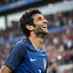 Diego Valeri, soccer star, flashing a big grin. Wearing blue jersey with Nike logo. Background of a crowded stadium is blurred.