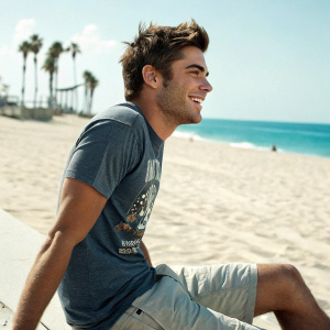 Zac Efron at the beach, wearing casual shorts and a graphic tee, with a relaxed smile.