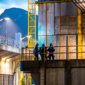 A promotional photo for Tom Clancy's Ghost Recon Breakpoint. Team of soldiers in dark gear standing near an old concrete building with ladders. Mountainous landscape in the background. Warm lighting casting shadows, creating a dramatic scene.