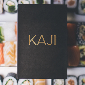 Kaji, a sushi restaurant logo, featuring a sleek black and gold design. Set against a backdrop showcasing an array of colorful, artistic sushi creations.