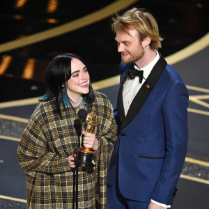 Billie Eilish and Finneas at the Academy Awards. Billie in a plaid outfit, holding an Oscar. Finneas in a blue suit. They stand on a stage with gold accents